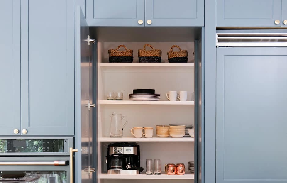 Blue kitchen cabinets with an open shelf displaying baskets, plates, mugs, and a coffee maker