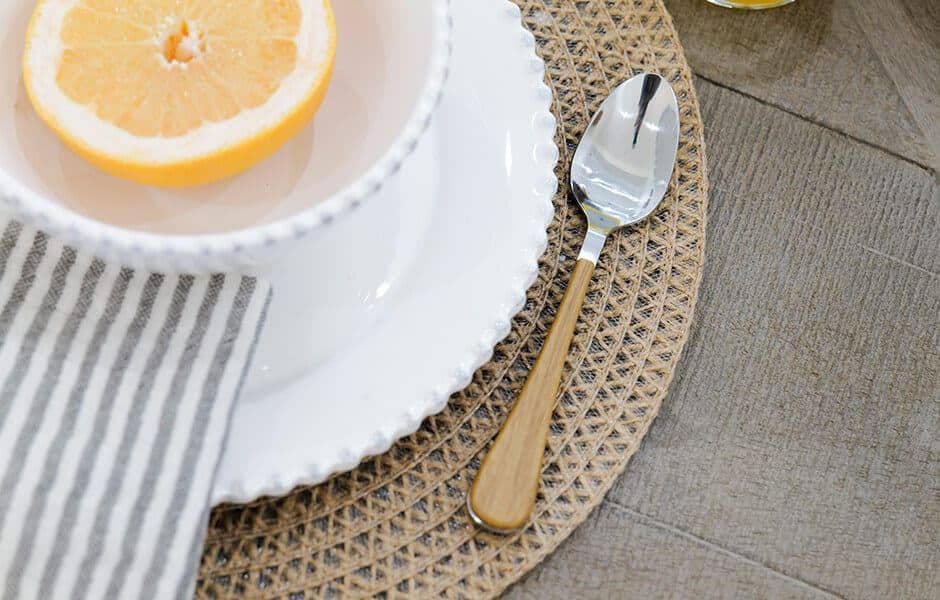 Table setting with a white plate, striped napkin, wooden-handle spoon, and a halved grapefruit on a woven placemat