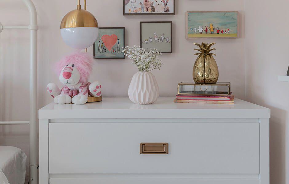 A child's bedroom with a white dresser, gold handle, stuffed lion, vase, pineapple, books, framed pictures, and a lamp