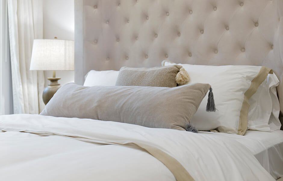 Double bed with neutral-colored pillows, featuring a tufted beige headboard and soft lighting from a bedside lamp