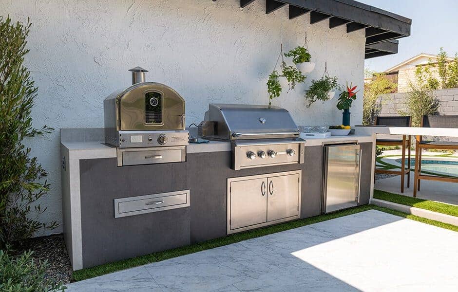 Outdoor kitchen with built-in grill, pizza oven, and mini-fridge near a poolside patio